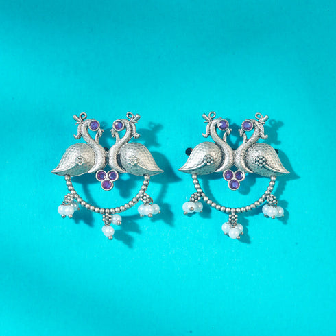 Elegant oxidized silver earrings featuring twin peacocks with ruby red stone accents and delicate pearl drops, perfect for traditional or festive occasions. Trenzify Store