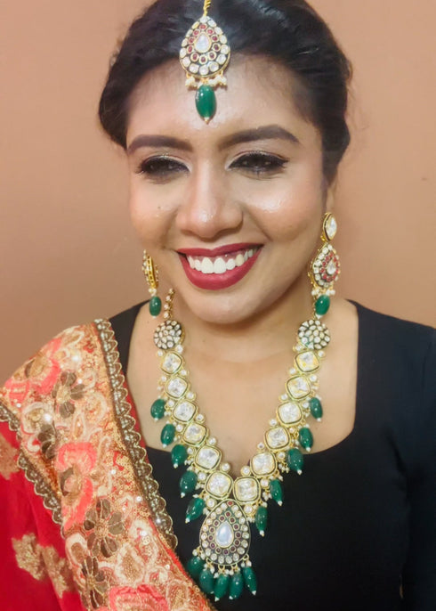 Model wearing gold plated Kundan necklace set with green stones, red enamel accents, pearls and matching earrings and maang tikka – traditional bridal jewelry from Trenzify Store.