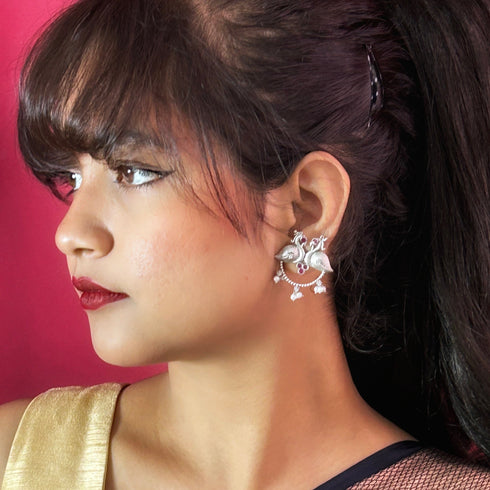 Elegant oxidized silver earrings featuring twin peacocks with ruby red stone accents and delicate pearl drops, perfect for traditional or festive occasions. Trenzify Store
